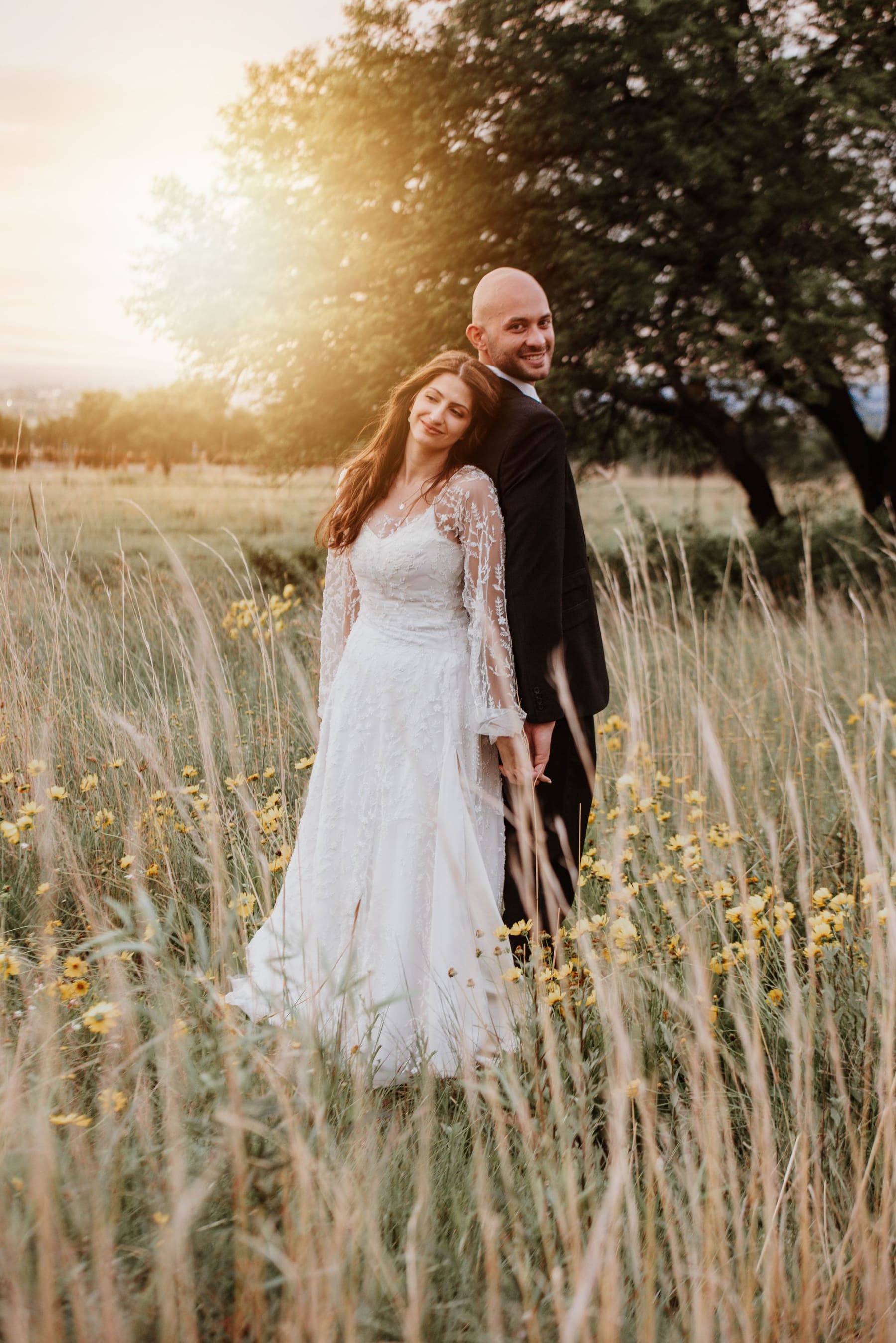 Um casal de noivos sorrindo num campo de flores silvestres, ao entardecer.