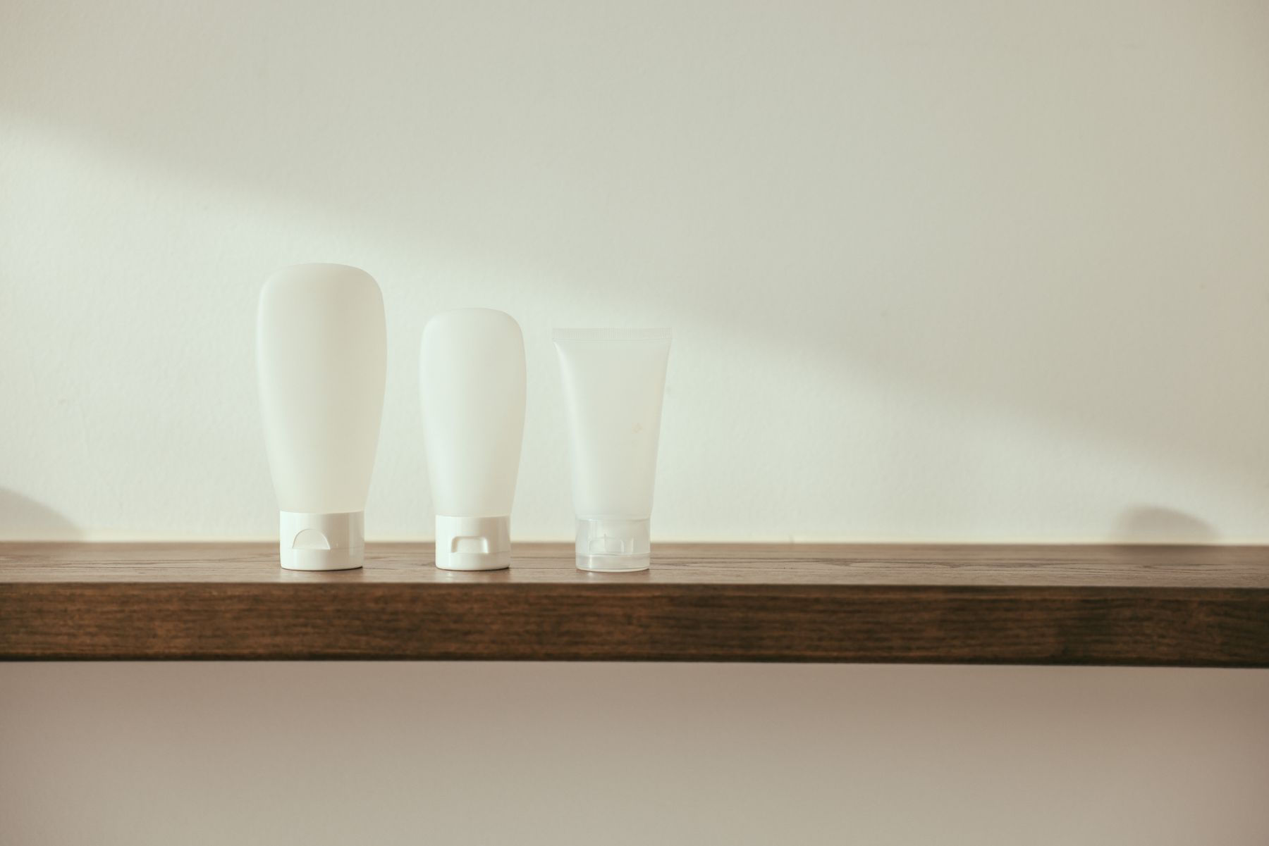Três frascos de skincare sem rótulo, em tons neutros, sobre uma prateleira de madeira.