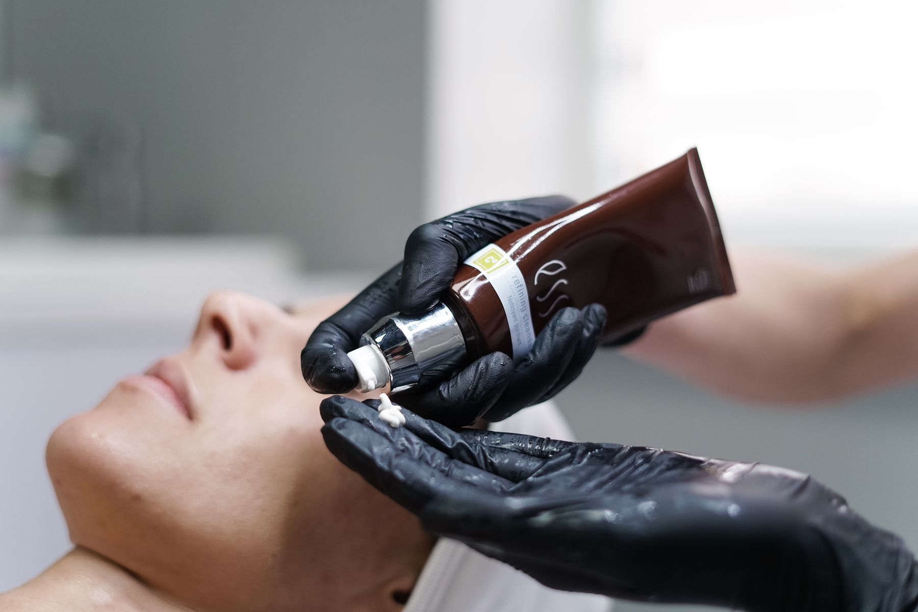 Mãos com luvas de nitrila aplicando um protocolo facial numa cabine de luz baixa, sob o foco de um aparelho.