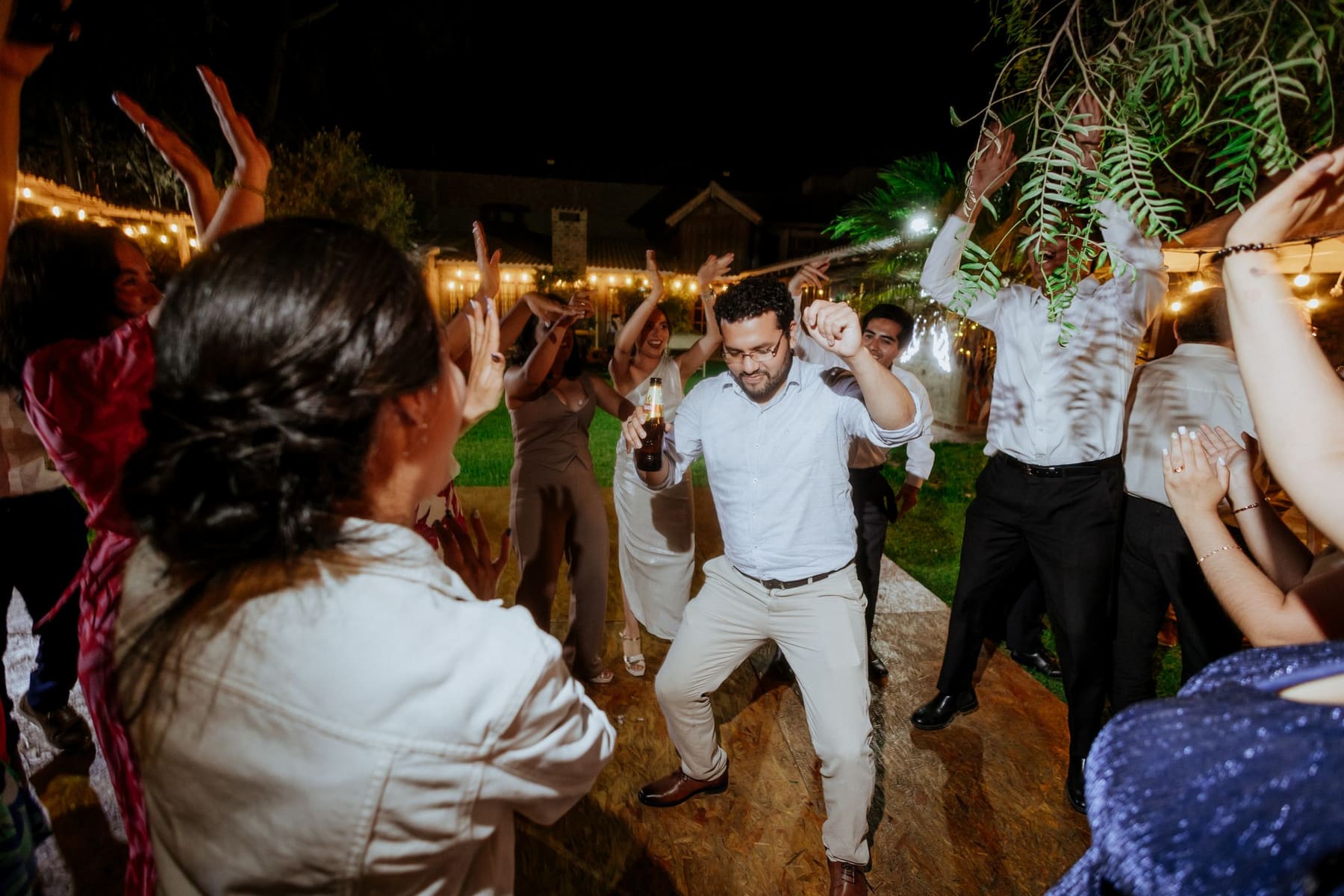 Convidados dançando na festa, à noite, sob luz quente de varal de lâmpadas.