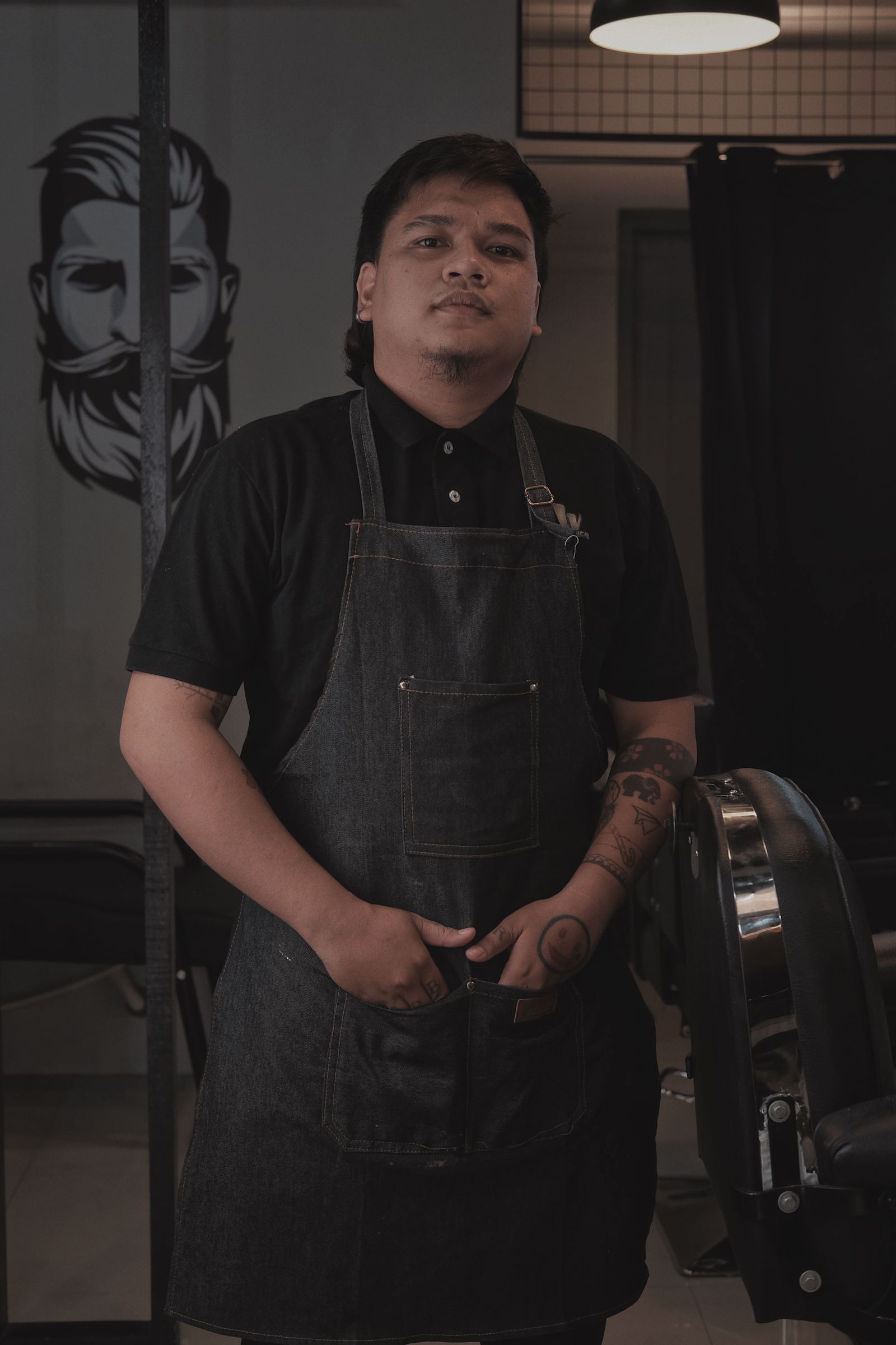 Téo Vidal, de avental de trabalho, em pé na barbearia, olhando para a câmera.