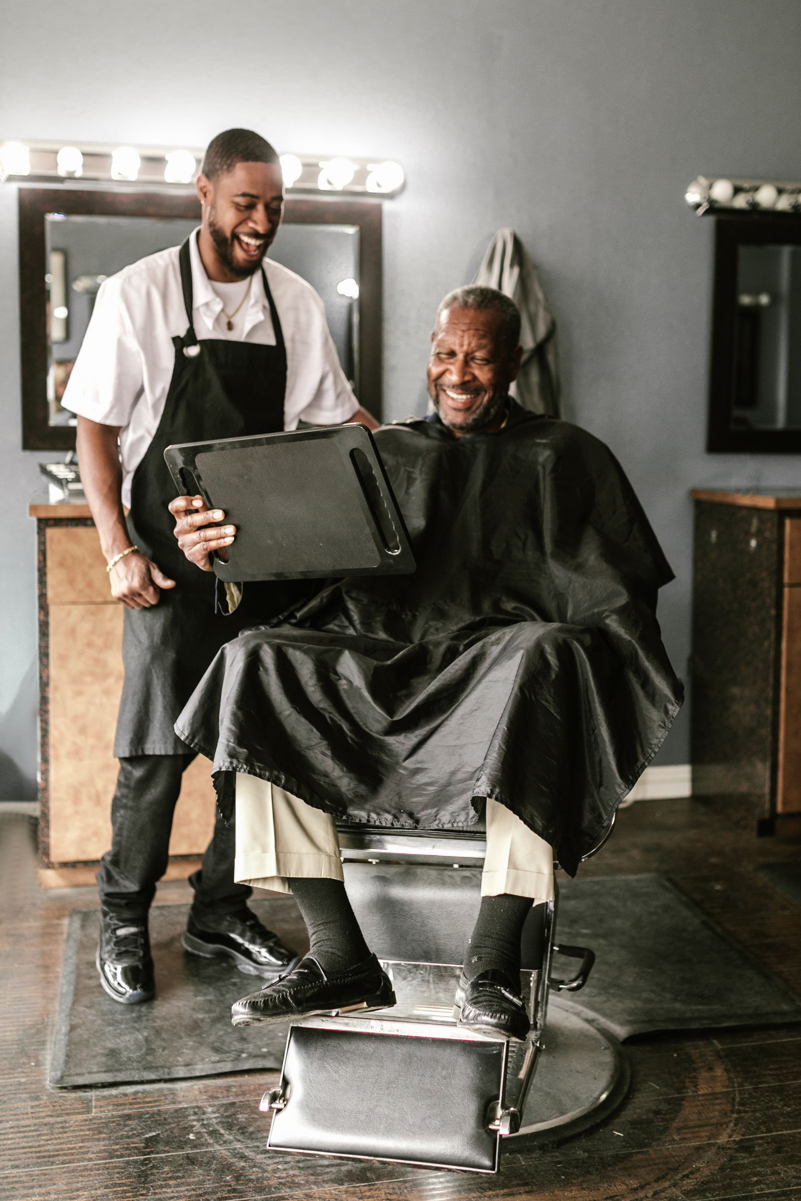 Barbeiro mostra o resultado do corte para o cliente com um espelho de mão, os dois sorrindo.